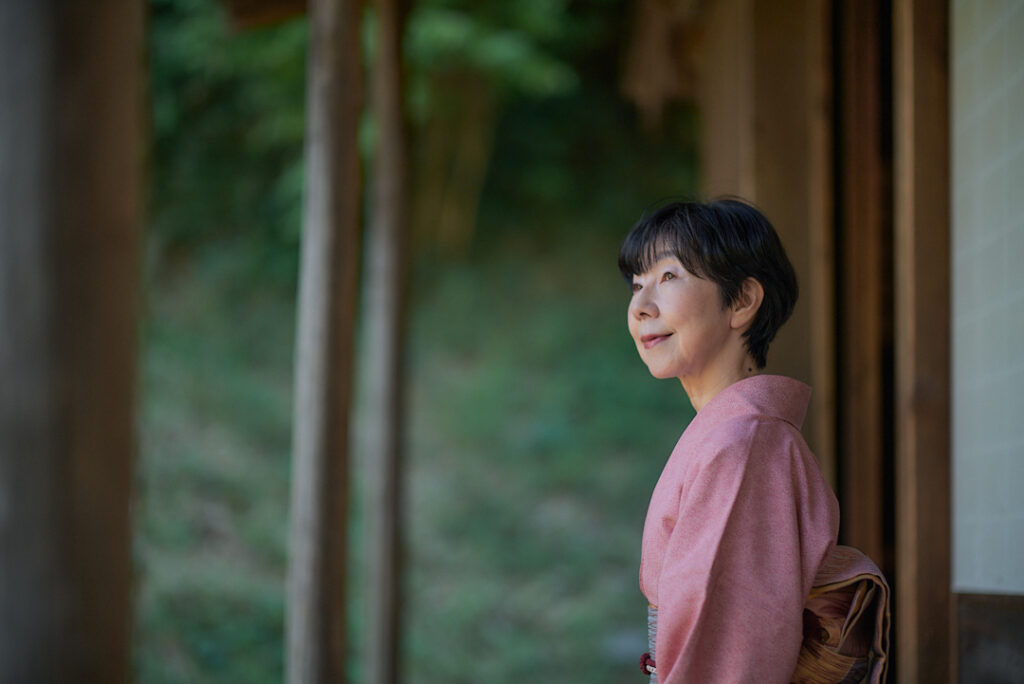 赤い着物を着て微笑む随筆家の石川真理子。日本家屋の縁側で凜としたたたずまい。