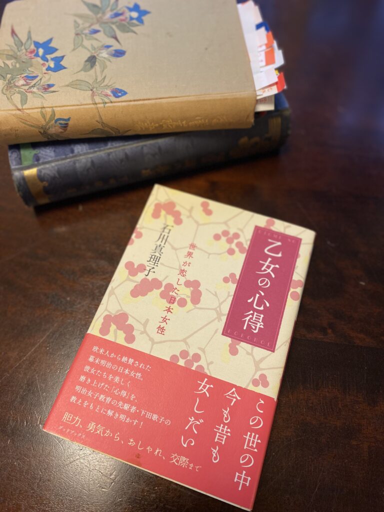 下田歌子の教えを読み解いた石川真理子の著作『乙女の心得』。ピンク色の帯が美しい。背後には明治時代の下田歌子の本が2冊重なっている。