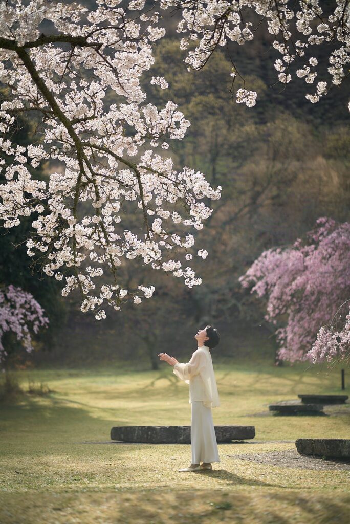 春の光が差し込むのどかな公園で、満開のソメイヨシノの大きな枝の下に立つ石川真理子。彼女は白く柔らかなブラウスとロングスカートを着用し、両手を少し前へ差し出すような仕草で、はらはらと舞い散る桜の花びらを愛おしむように見上げている。背景には、ピンク色の枝垂れ桜や、木々の優しい緑が広がり、清らかで美しい瞬間を捉えている。
