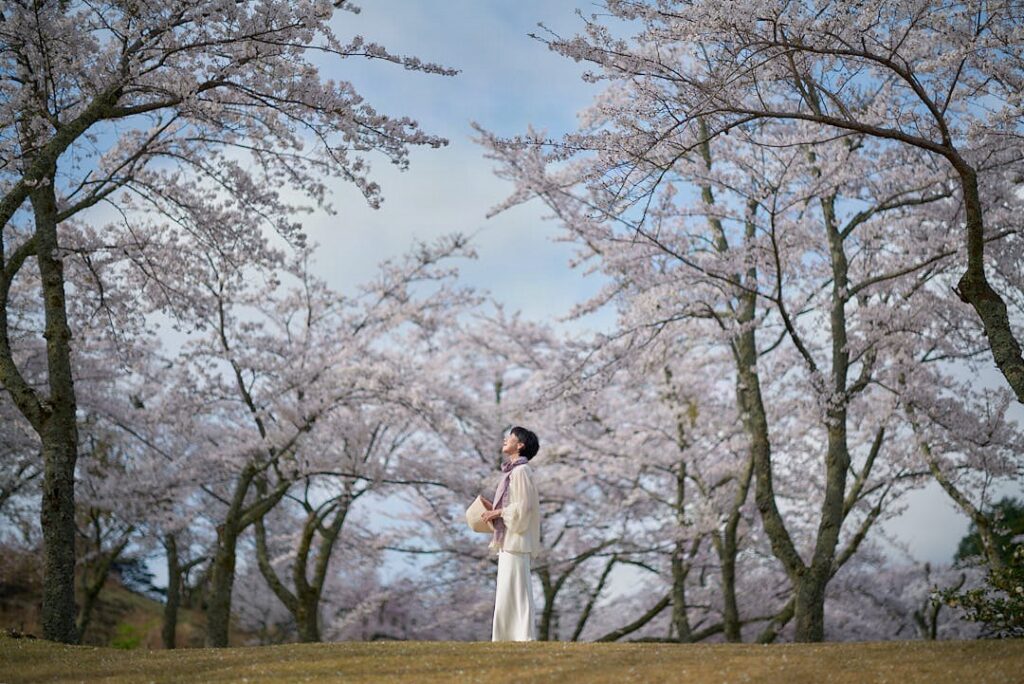 青空と、遠くまで続く満開の桜のトンネルを背景に、大地に一人立つ石川真理子の全身像。彼女は白いブラウスとロングスカートを着用し、左手に麦わら帽子、首元に紫色のストールを巻き、清々しい空気の中で、桜に囲まれた空を見上げている。左右の桜の木が、彼女を包み込むようなフレームとなり、広大な自然の中に身を置く、凛とした一瞬が捉えられている。