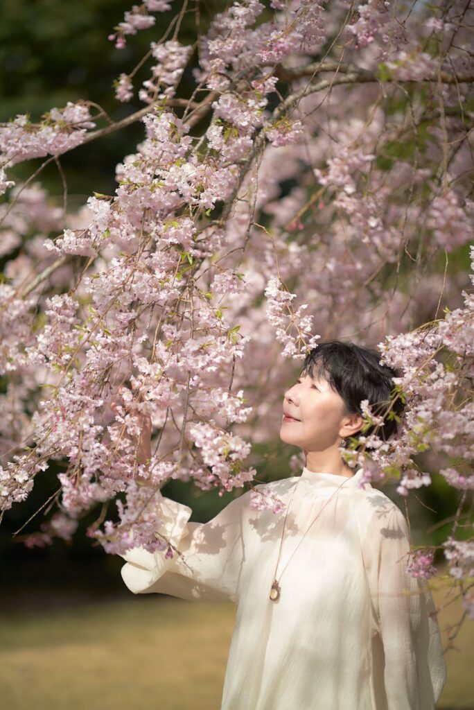 満開の枝垂れ桜が、薄ピンク色のカーテンのように垂れ下がる中、その傍らに立つ石川真理子。彼女は白いブラウス姿で、自身の頭上に優しくかかる桜の枝を見つめている。彼女の視線と、流れるような桜の枝が交差する構成は、人と自然の調和を感じさせる。光が滲むような柔らかさと、繊細な階調が、彼女の柔らかな表情と桜の美しさを際立たせている。