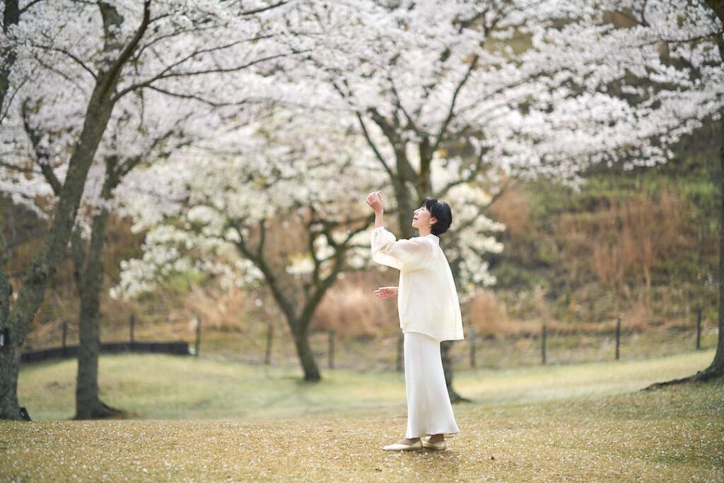 満開の桜の木の下で、白いブラウスとロングスカート姿の石川真理子が立ち、右手を高く掲げて、舞い落ちてくる桜の花びらを受け止めようとしている。彼女は優しい表情でその手を見つめており、背景には桜の森が白く霞むように広がっている。ライカ・タンバールレンズがもたらす、光が飽和したような白く柔らかい描写が、彼女の周囲の空気を清らかで神秘的なものにしている。