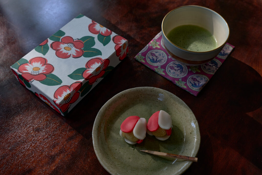 若草色のお皿に盛られた和菓子の「のりこぼし」。紅い椿の絵柄が描かれた萬々堂通則の箱と、一碗の抹茶が添えられている。