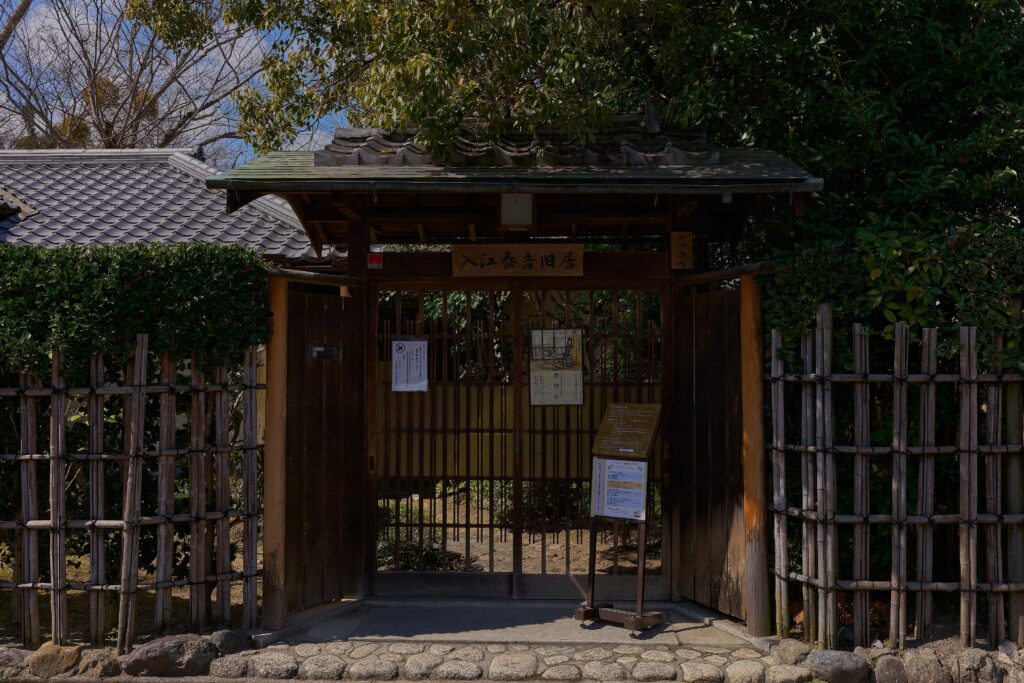 入江泰吉旧宅の門構え。昔ながらの日本風の門、両脇には垣根がある。門の向こうには前庭があり、瓦屋根が見えている。