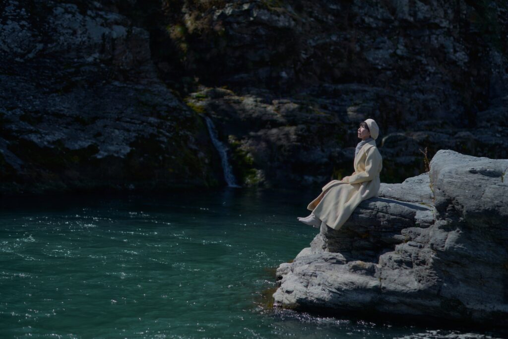 青く澄んだ川辺の巨石の上で寛ぐ石川真理子。白いベレー帽と白いコート姿でくつろいでいる。吉野宮遺跡近くの「夢のわだ」で魚住心が撮影。