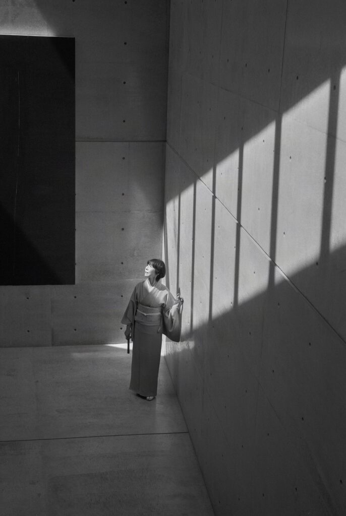 コンクリートの無機質な空間に鋭い斜めの光と影が落ちる中、和服姿で凛と佇む石川真理子の全身モノクロ写真。冷徹な世界の中で自らの軸と美意識を保つ姿を象徴している。