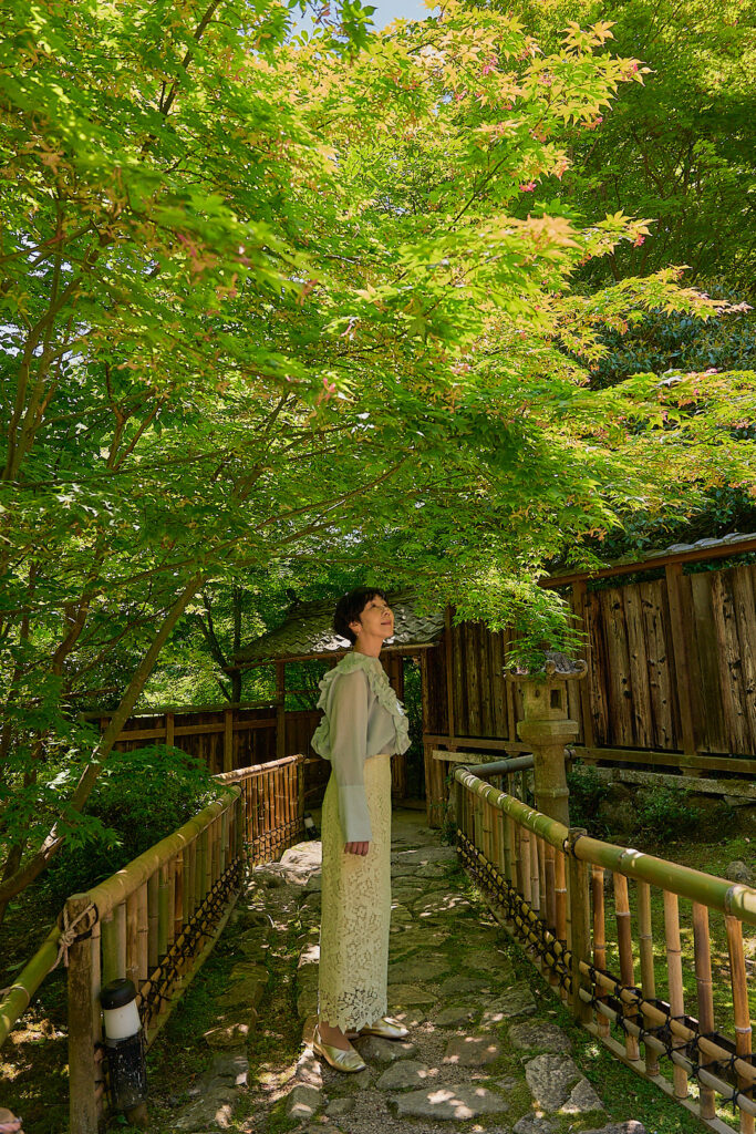 柔らかな新緑の下、水色のフリルブラウスと白いレーススカートを纏った石川真理子が、優しく微笑みながら木々を見上げている。