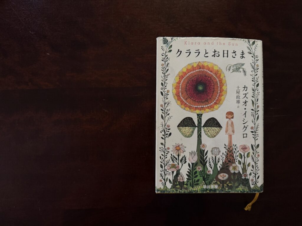 深い焦げ茶色の木目の机の上に置かれた、カズオ・イシグロ著『クララとお日さま』の日本版単行本（早川書房）。表紙には大きなひまわりのようなお日さまと、その傍らに佇む少女、そして周囲を囲む草花の美しいイラストが描かれている。