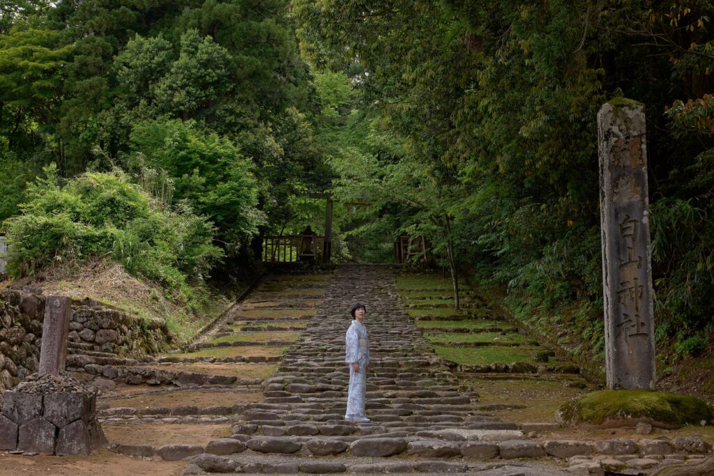 福井県の古刹・平泉寺白山神社の石段前に佇む石川真理子。水色の古典柄の着物が苔と青紅葉に映えている。魚住心がライカで撮影。