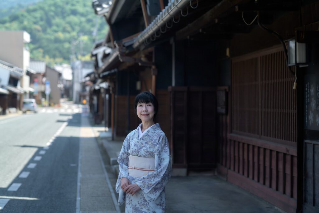 歴史的な木造建築が並ぶ日本の古い街並みを背景に、薄い青紫色に繊細な柄が描かれた着物を着た女性（石川真理子）が、自然な笑顔で立っているポートレート写真。魚住心 撮影