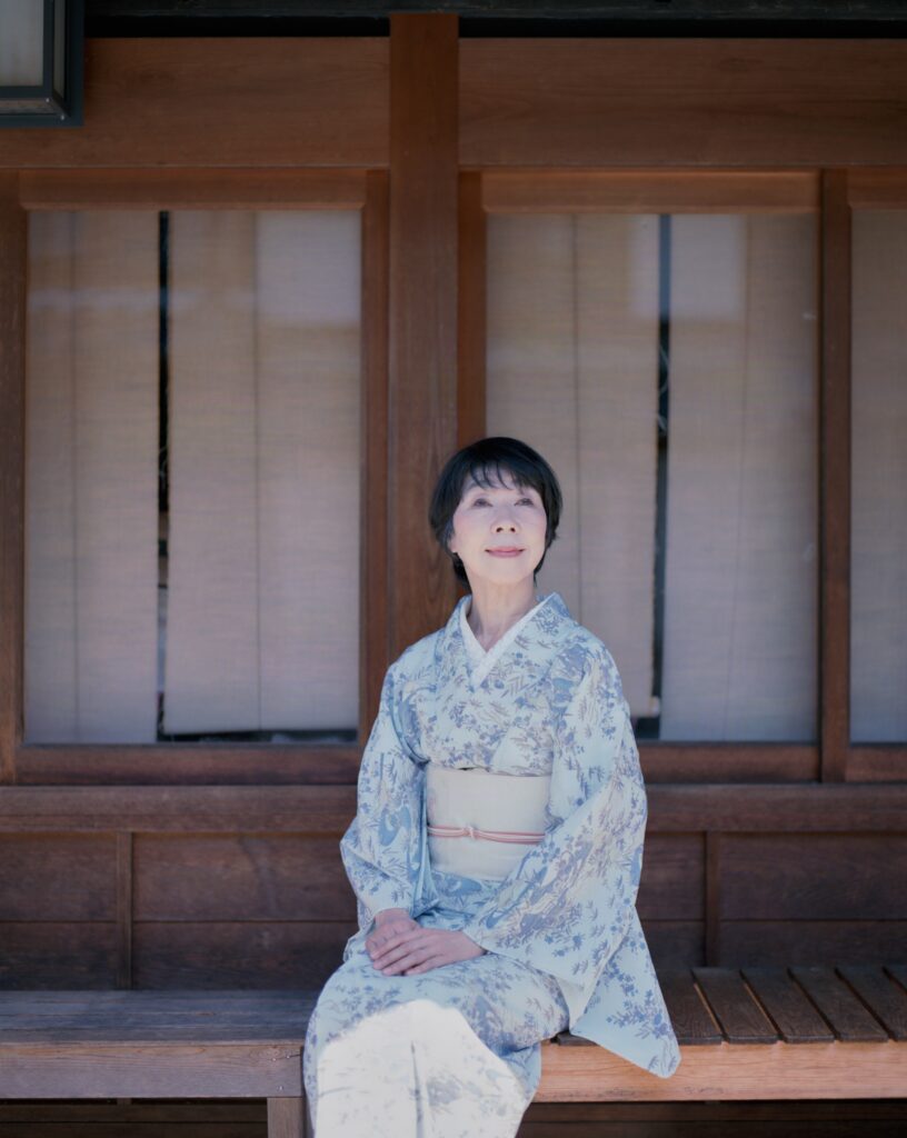 短い髪の日本人女性が、複雑な伝統模様の青い着物と白い帯を着用し、風化した木製のベンチに座って、伝統的な障子スクリーンの前で上を見上げて、穏やかで考え深い表情を浮かべている様子。