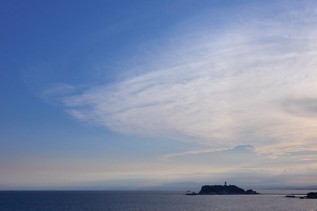 8月の夕暮れ、青い空と劇的な白い雲が広がる江ノ島沖の海原。遠くに江ノ島の島影が見える。