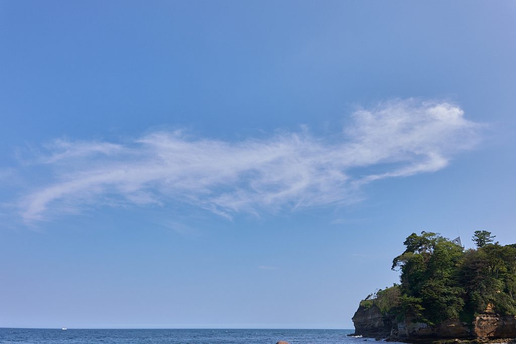 澄み切った青空に浮かぶ白く筋状の雲。下部には穏やかな海が広がり、右側には木々に覆われた岩の断崖が見える。