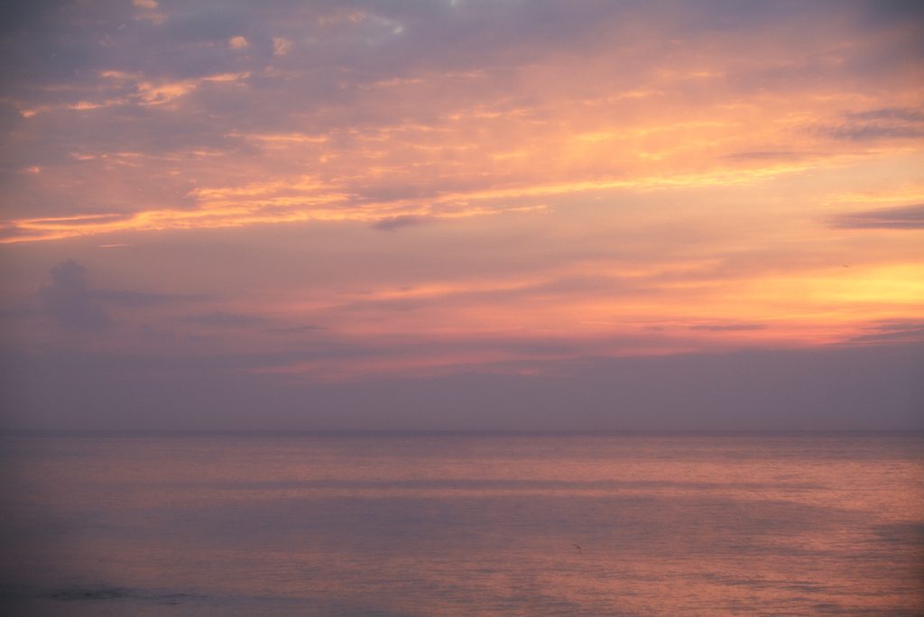 ピンクとオレンジ色に染まる夕暮れの海辺の空。静かな海面が空の色を反射し、遠くにぼんやりと雲が浮かんでいる。