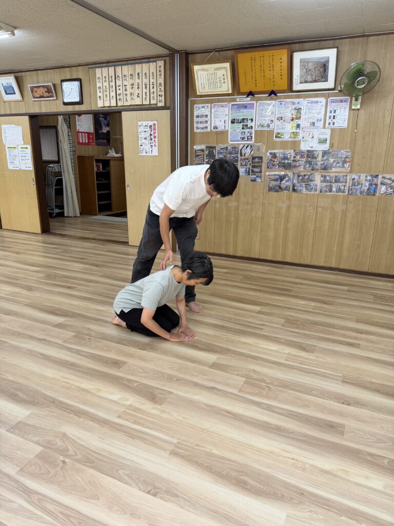 床に正座をして両手をつき、丁寧にお辞儀をしている女性。その背中を、小野川さんが上から手で押さえている様子。
