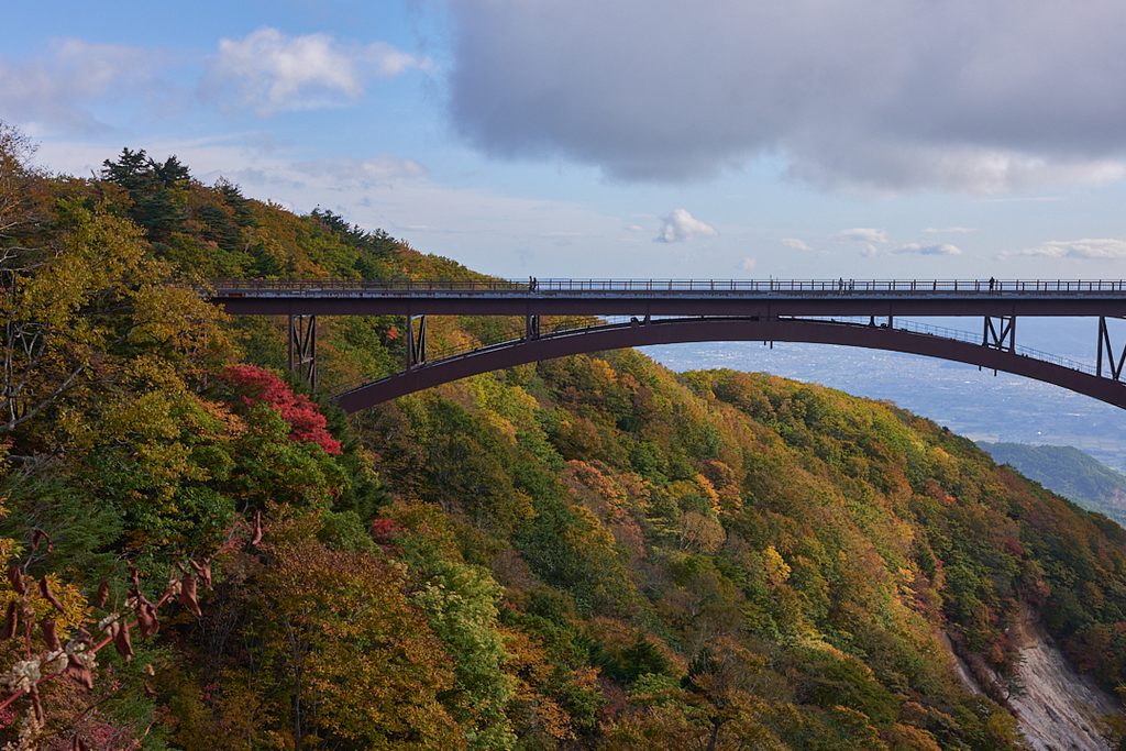 紅葉が始まった山腹をまたぐアーチ橋と、その向こうに遠く広がる平野と秋の空